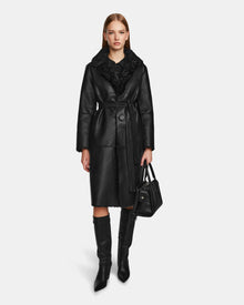 A woman stands facing forward, wearing the Rudsak ANNABELLE - L124544 Black shearling coat, knee-high black boots, and holding a black handbag. She has straight light brown hair, a neutral expression, and is set against a white background.