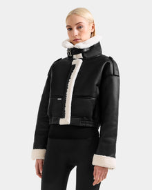 A woman with straight blonde hair wears the Rudsak APRIL - L125517 Black cropped jacket with white shearling trim, paired with black fitted pants, standing against a plain light gray background.