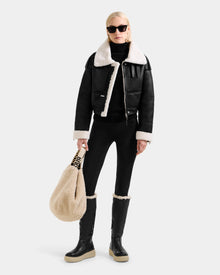 A woman in sunglasses wears Rudsak's APRIL - L125517 Black shearling-lined jacket with black leggings and black knee-high boots with cream accents. She holds a large beige faux-fur bag against a plain light gray background.