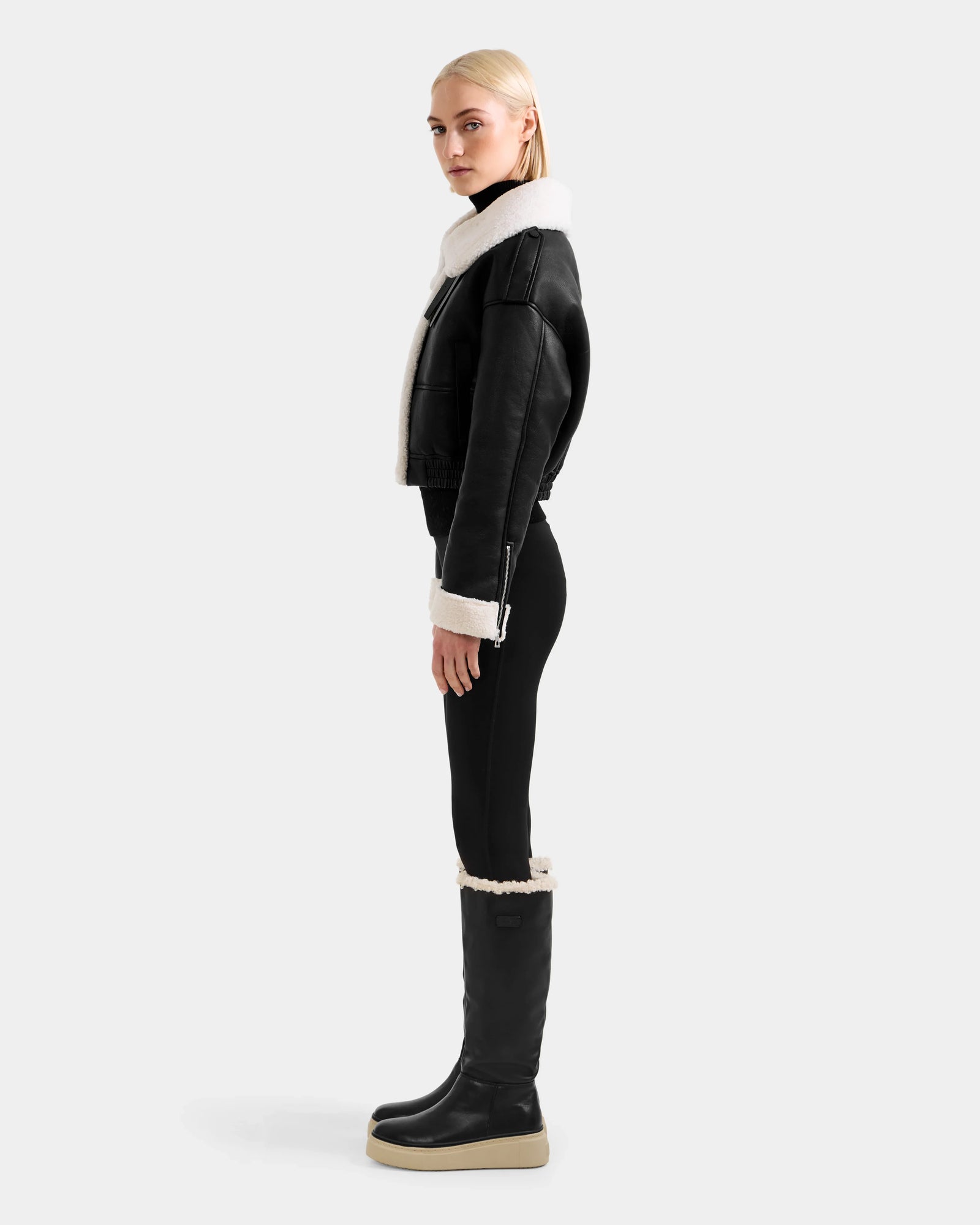 A woman with blonde hair stands in profile against a light gray background, wearing the Rudsak APRIL - L125517 Black shearling jacket, paired with black leggings and tall black platform shearling boots.