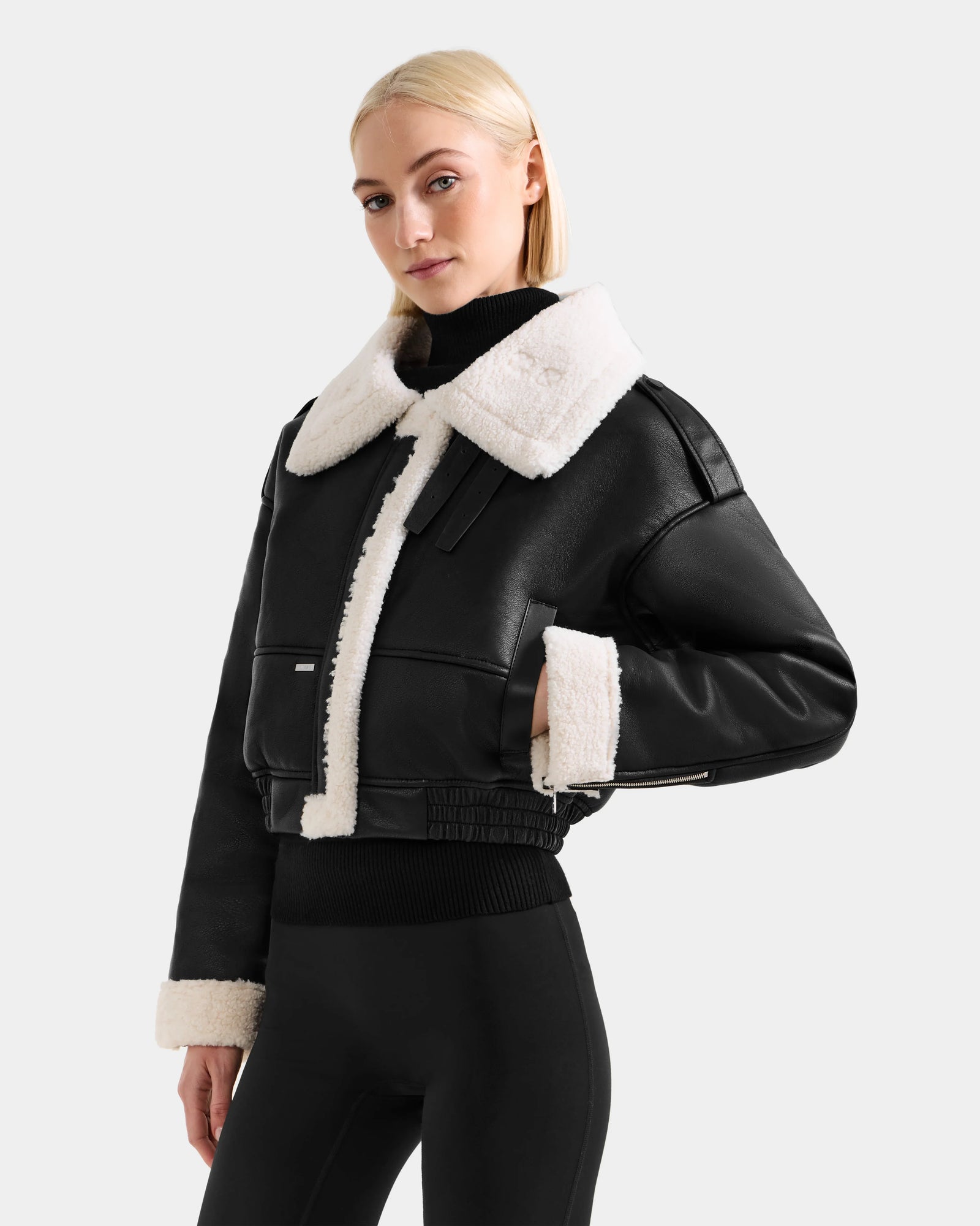 A woman wearing the Rudsak APRIL - L125517 Black vegan leather bomber with white faux shearling trim, styled with a black turtleneck and leggings, stands against a plain light background.
