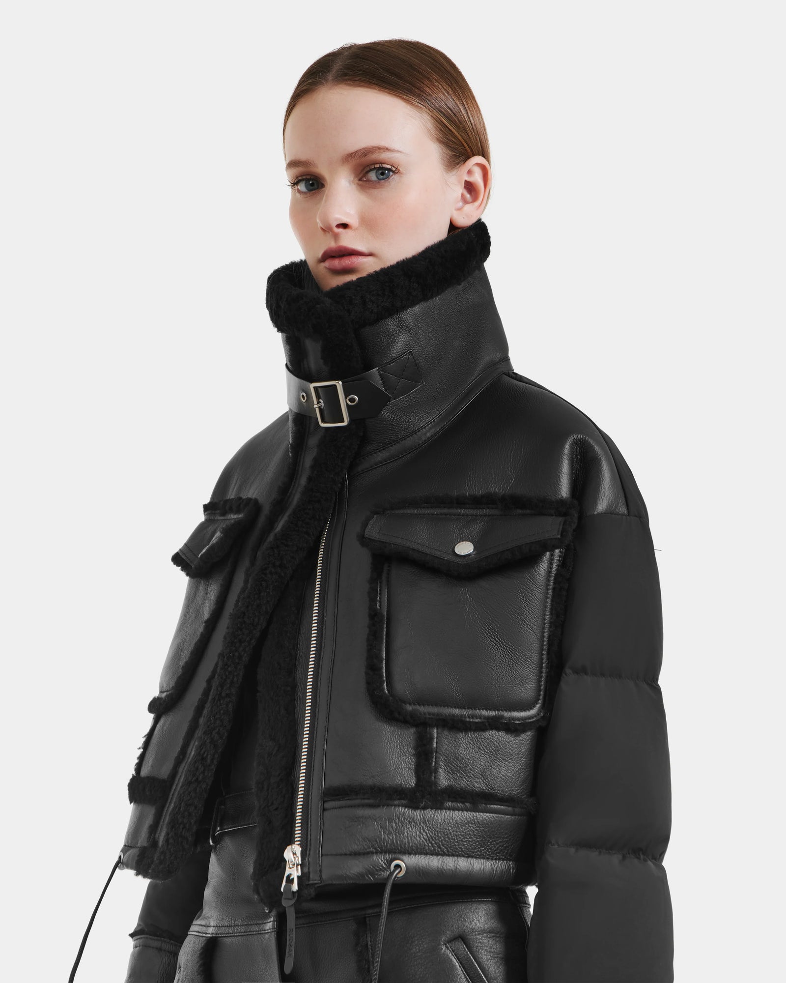 A woman wears the Rudsak ARIELLA - 8125505 Black cropped shearling puffer with a high collar, front zipper, large flap pockets, and a belted neck, standing against a white background—ideal for cold-weather style.