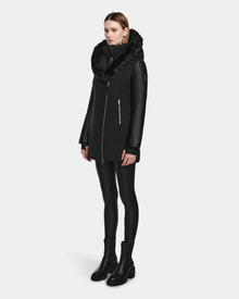 A woman wears the Rudsak AURELLE - L124538 Black womens down parka with a fur-lined hood, vegan leather sleeves, black leggings, and black ankle boots against a plain light gray background.
