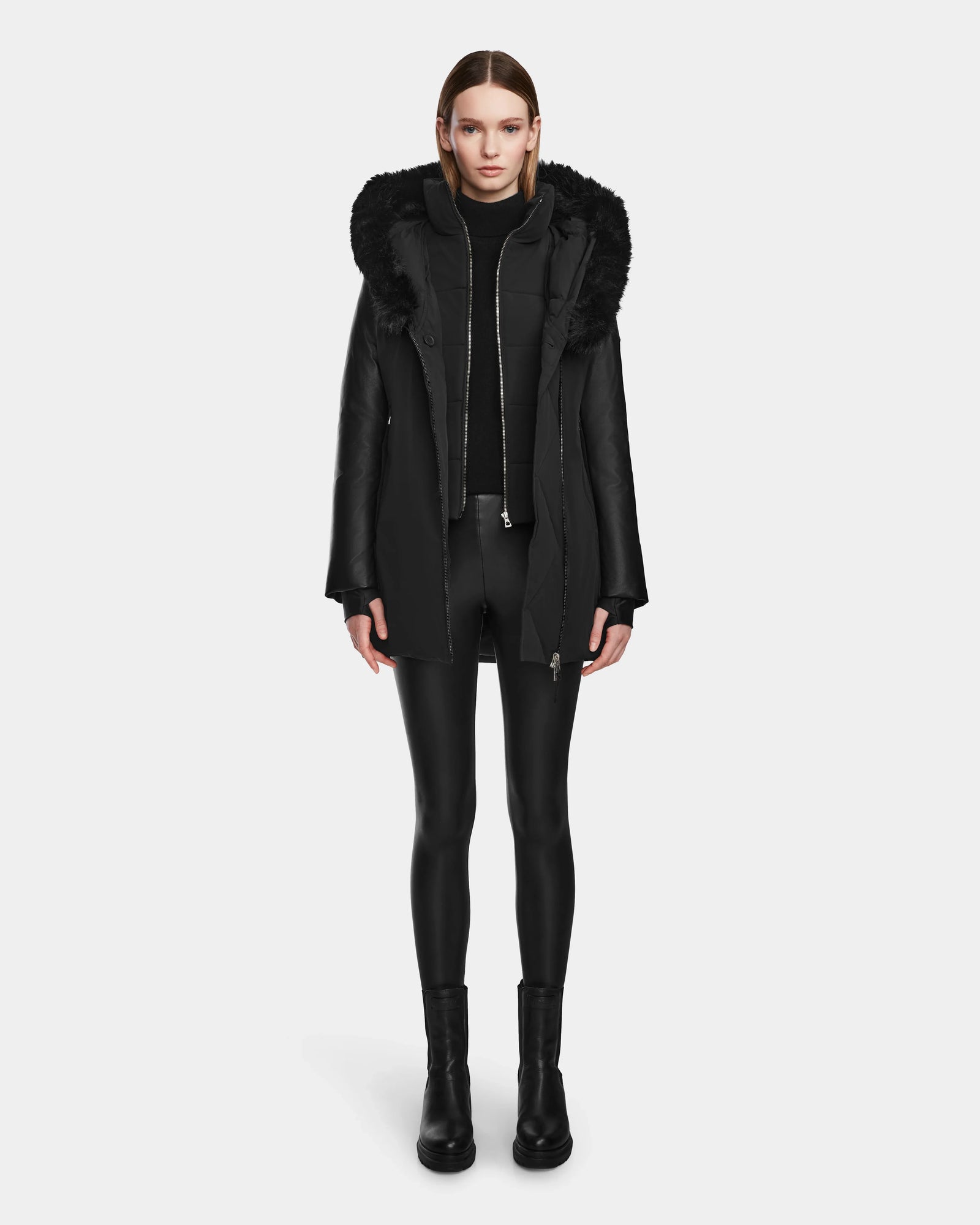 A woman faces forward wearing the Rudsak AURELLE - L124538 Black womens down parka with vegan leather sleeves and a fur-lined hood, styled with a black turtleneck, leggings, and boots against a plain white background.