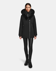 A woman stands facing forward wearing the Rudsak AURORE - L123561 Black/Black women’s short fitted down parka with faux fur trim, paired with black leggings and black ankle boots, against a plain white background.