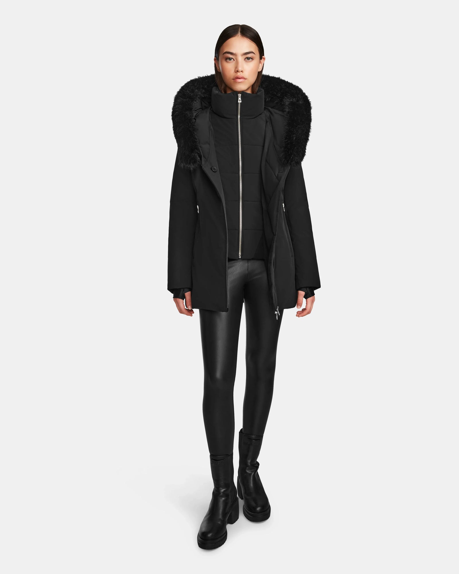 A woman wears the Rudsak AURORE - L123561 Black/Black women's short fitted down parka with a large faux fur-trimmed hood, paired with black leggings and boots, standing against a plain white background.