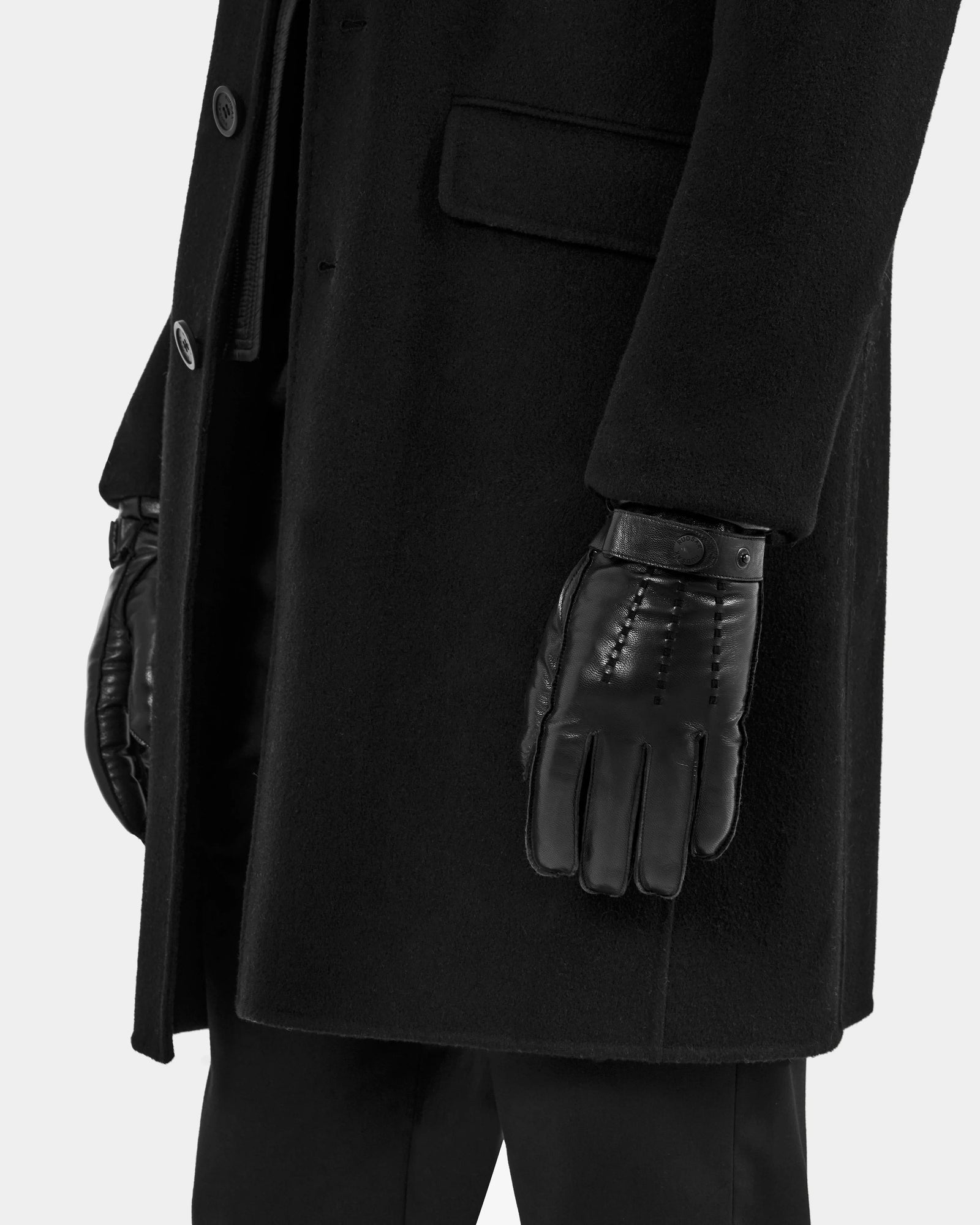 A person wears Rudsak's BORIS - M425001 Black coat, black pants, and men’s leather gloves with wool-cashmere lining, standing with their arm by their side against a plain light background—a true cold-weather essential.