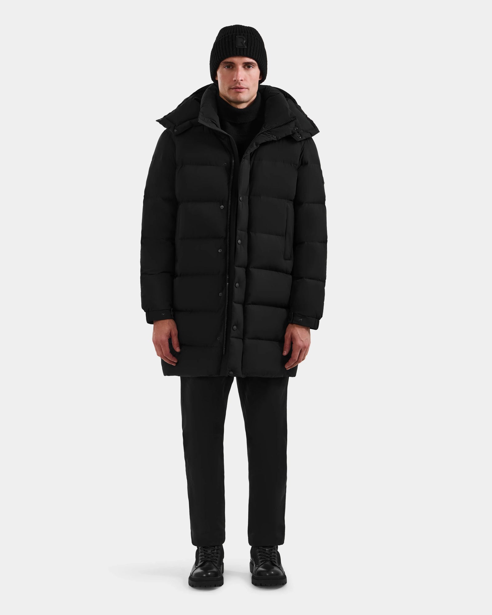 A person stands facing forward, wearing the Rudsak BRAYDEN M-M - M125535 Black men’s down puffer coat, paired with a black knit beanie, black pants, and black shoes against a plain white background.