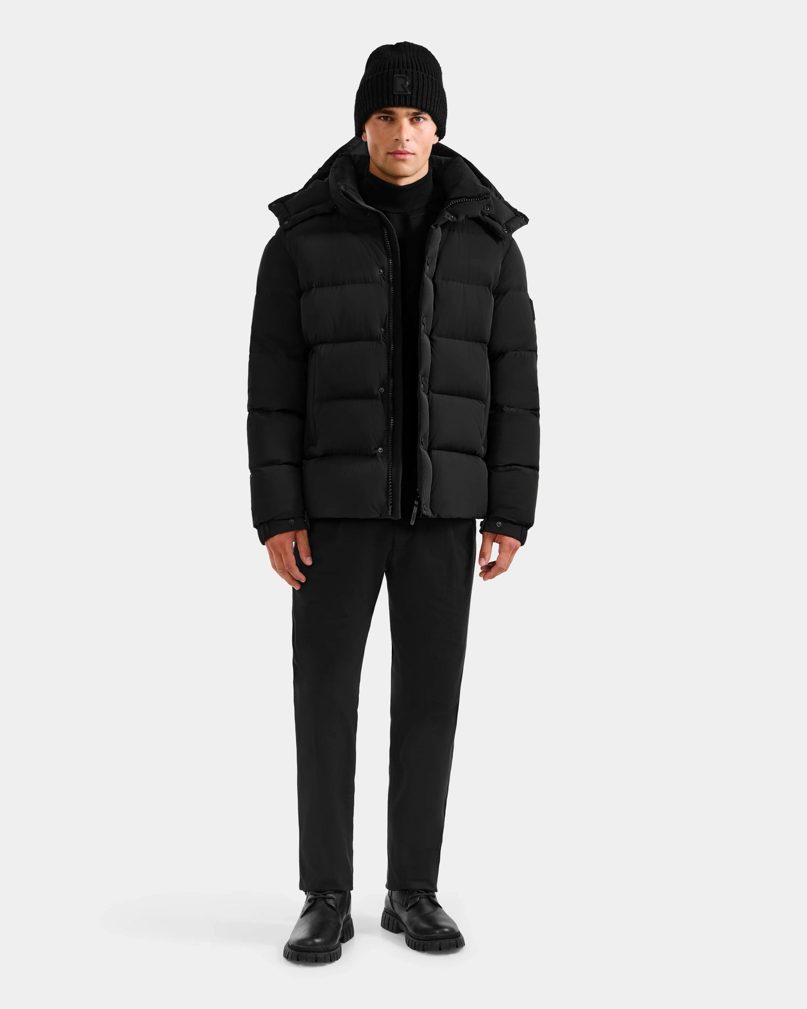 A person faces forward wearing the Rudsak BRAYDEN M-S - M125524 Black men’s down puffer jacket, paired with a black beanie, pants, and shoes against a plain light background.