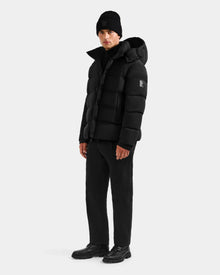 A man wearing the Rudsak BRAYDEN M-S - M125524 Black puffer jacket, black knit beanie, black pants, and black shoes stands against a plain light gray background.