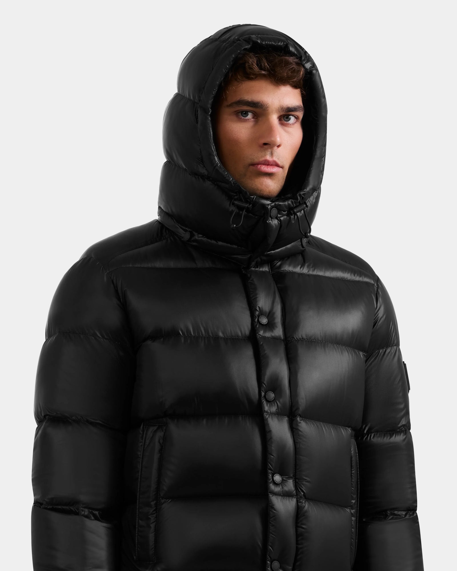 A man wearing the Rudsak BRAYDEN S - M125525 Black quilted down puffer with the hood up, stands facing forward against a plain light gray background.