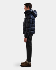 A man stands in profile against a plain white background wearing the Rudsak BRAYDEN S - M125525 Navy quilted down puffer jacket with a hood, paired with dark cuffed pants and brown suede boots.