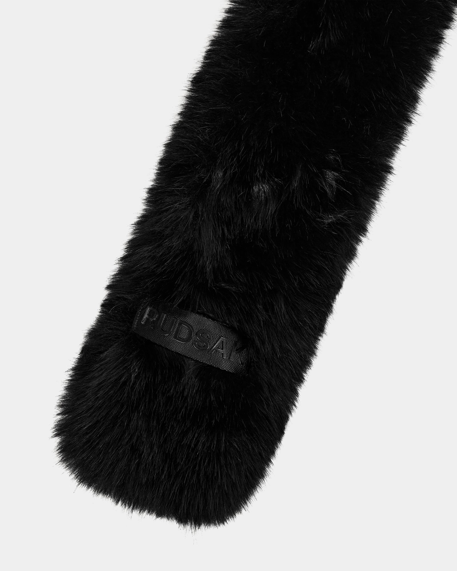 A close-up of the Rudsak CARMY - L425534 Black women’s faux-fur scarf, highlighted by a rectangular embossed patch, showcases its plush warmth against a plain light gray background.