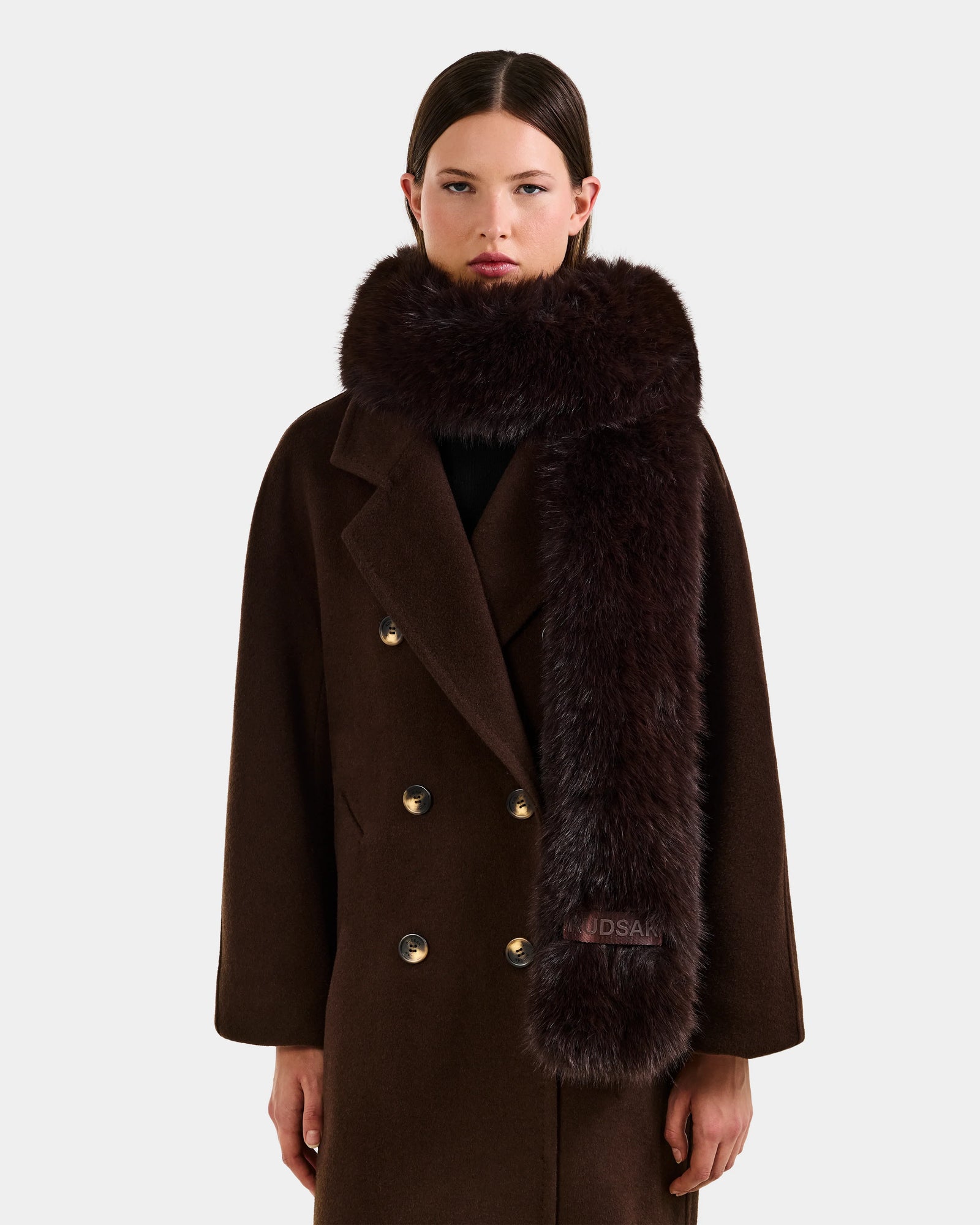 A woman wears the CARMY - L425534 Espresso coat by Rudsak with a thick faux-fur scarf, standing against a plain light background—a stylish and cozy choice for cold weather.