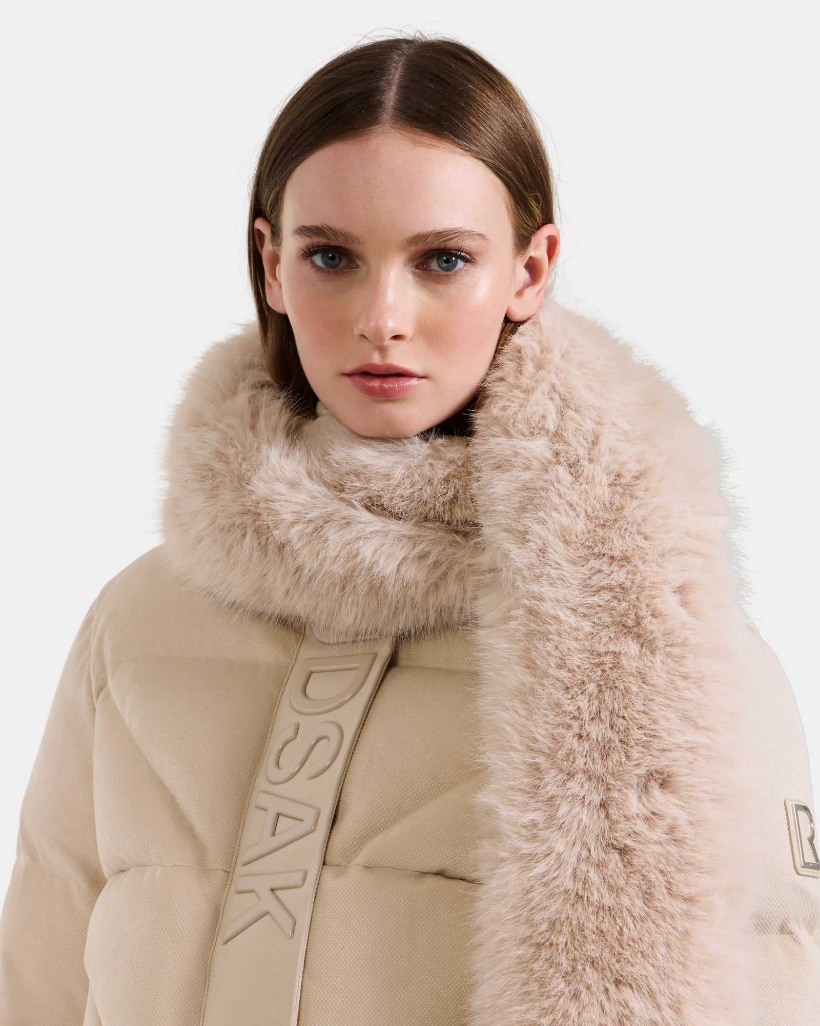A woman with straight brown hair wears the Rudsak CARMY - L425534 Vanilla puffer coat with a plush fur collar, standing with a serious expression against a plain light background.