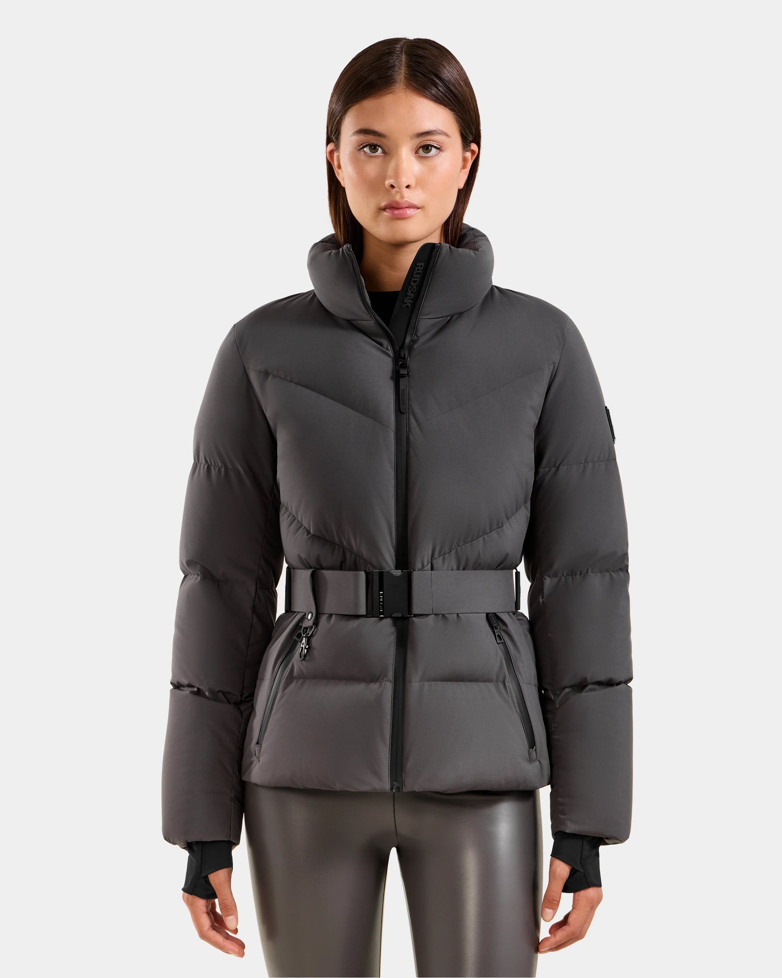 A woman stands facing forward, modeling the Rudsak CHRISTELLE M - L125542 Grey jacket—a dark gray, weather-resistant down puffer with a belted waist and zippered pockets—against a plain white background.
