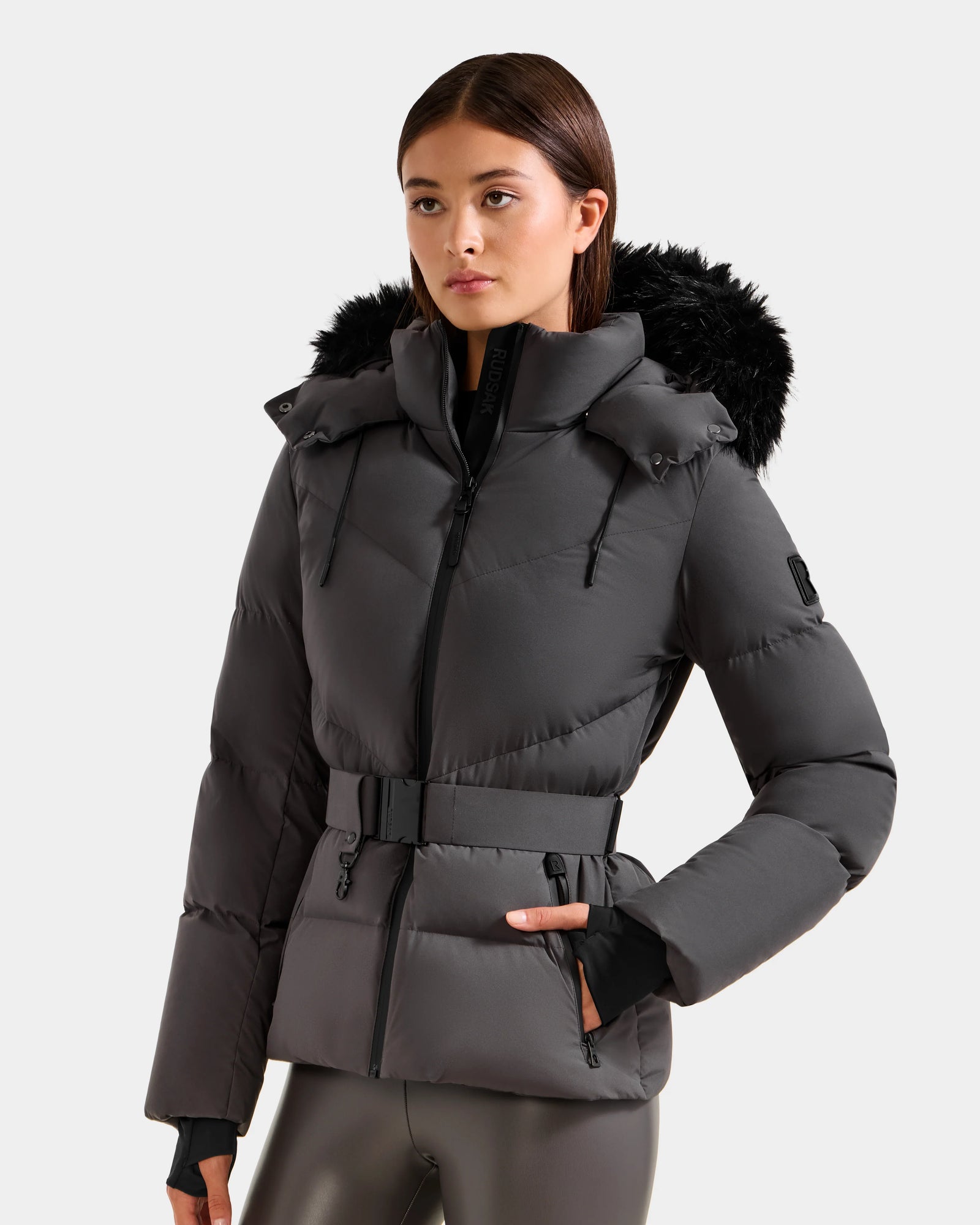 A woman models the Rudsak CHRISTELLE M - L125542 Grey jacket, featuring a dark gray weather-resistant design, black faux fur hood trim, belted waist, and zip-up front, posing with one hand in her pocket.
