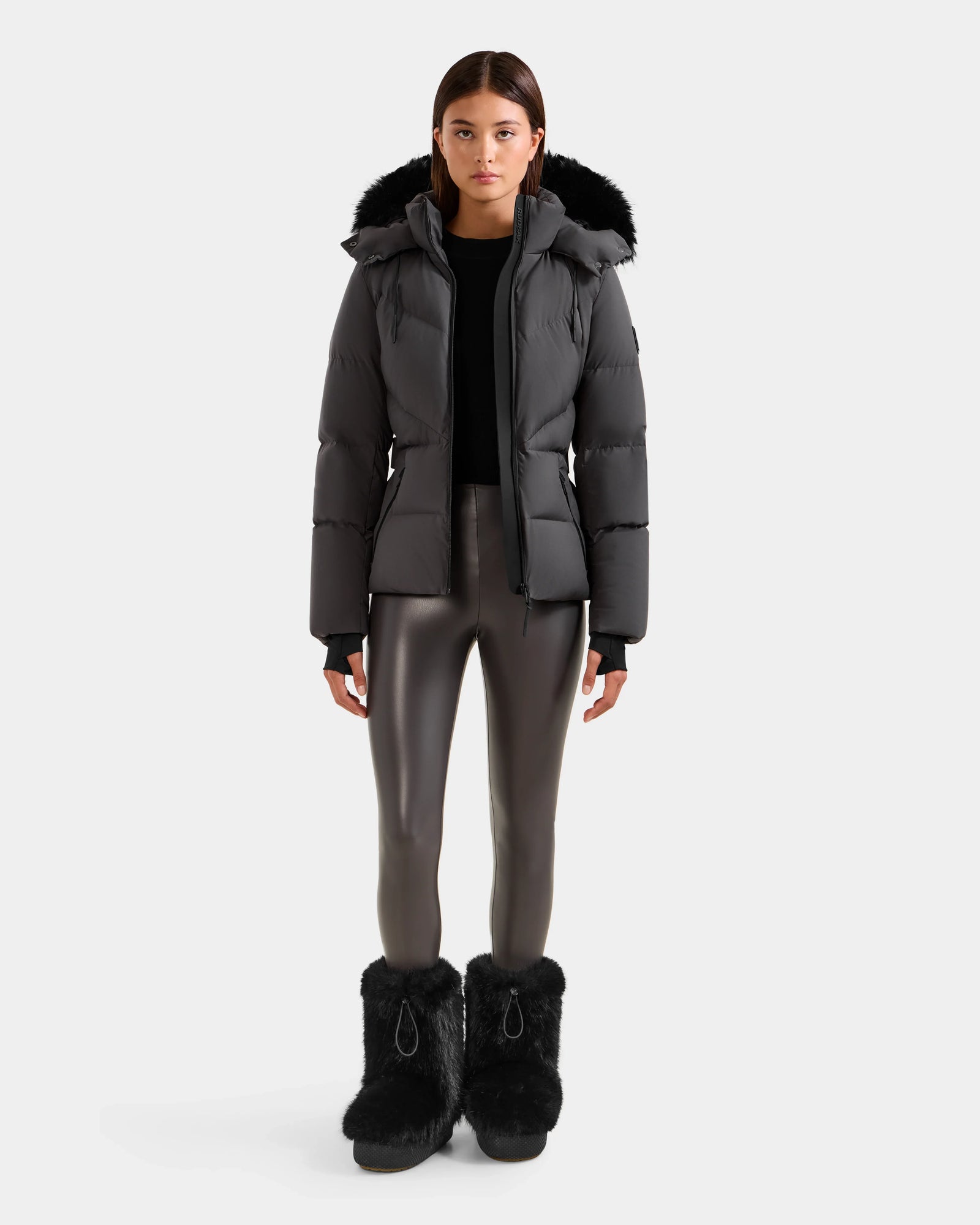 A woman faces forward wearing the Rudsak CHRISTELLE M - L125542 Grey quilted down puffer jacket with a fur-trimmed hood, shiny gray leggings, and black fur boots against a plain light background.