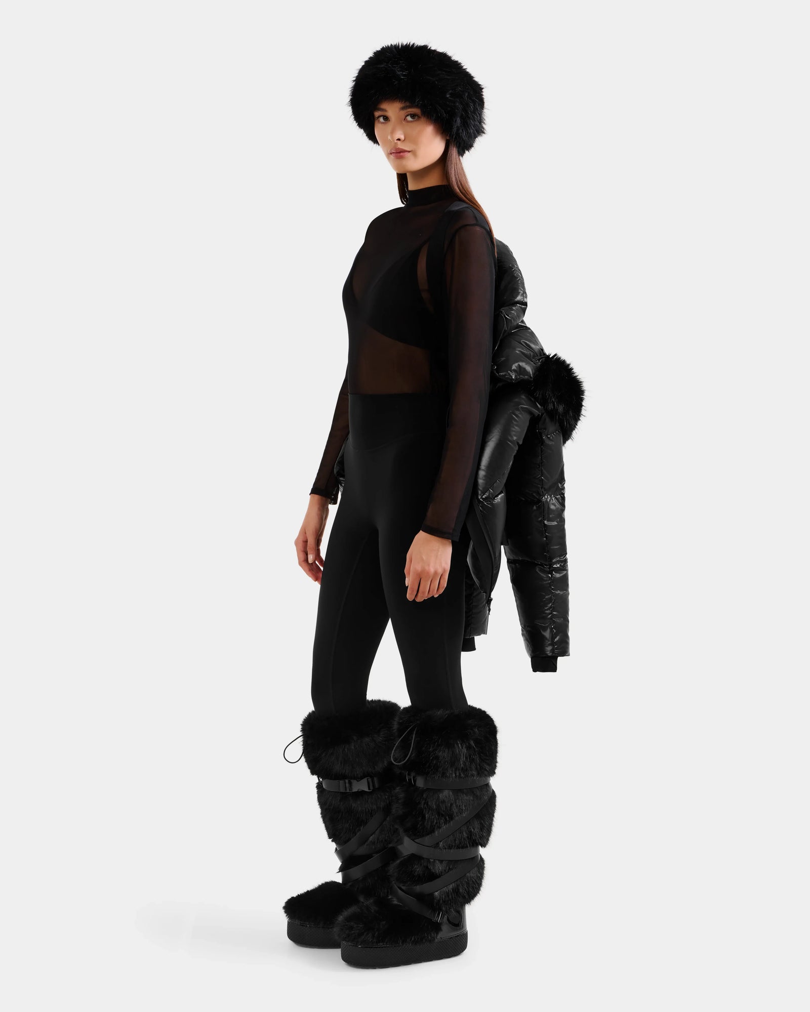 A woman stands against a light background holding the Rudsak CHRISTELLE S - L125544 Black women’s down puffer jacket with faux fur trim over her shoulder, styled with black fur boots, leggings, a sheer top, and a fur hat.