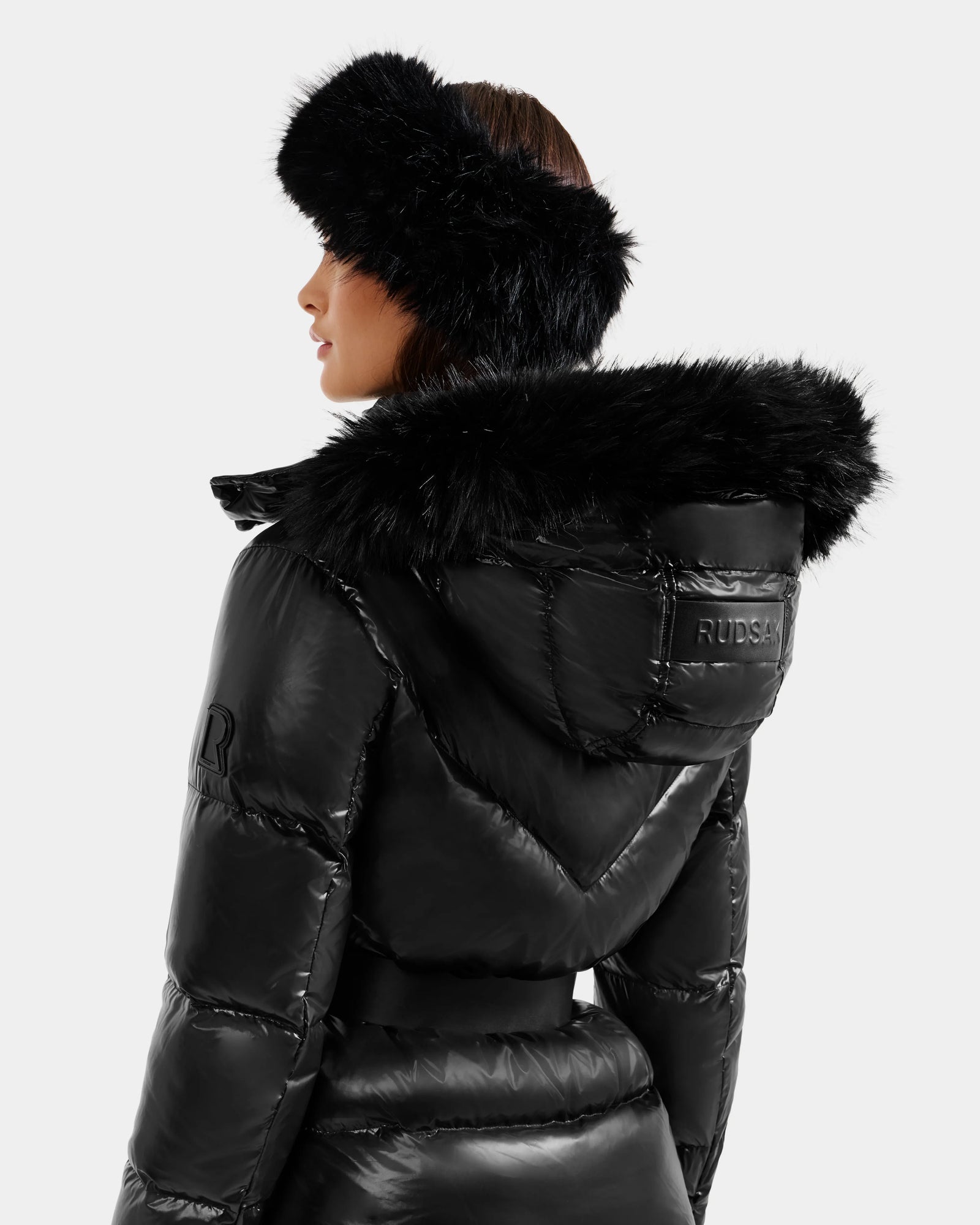 A woman wearing the Rudsak CHRISTELLE S - L125544 Black down puffer jacket with a faux fur-trimmed hood and a matching black fur headband stands against a plain light background, facing partly away from the camera.