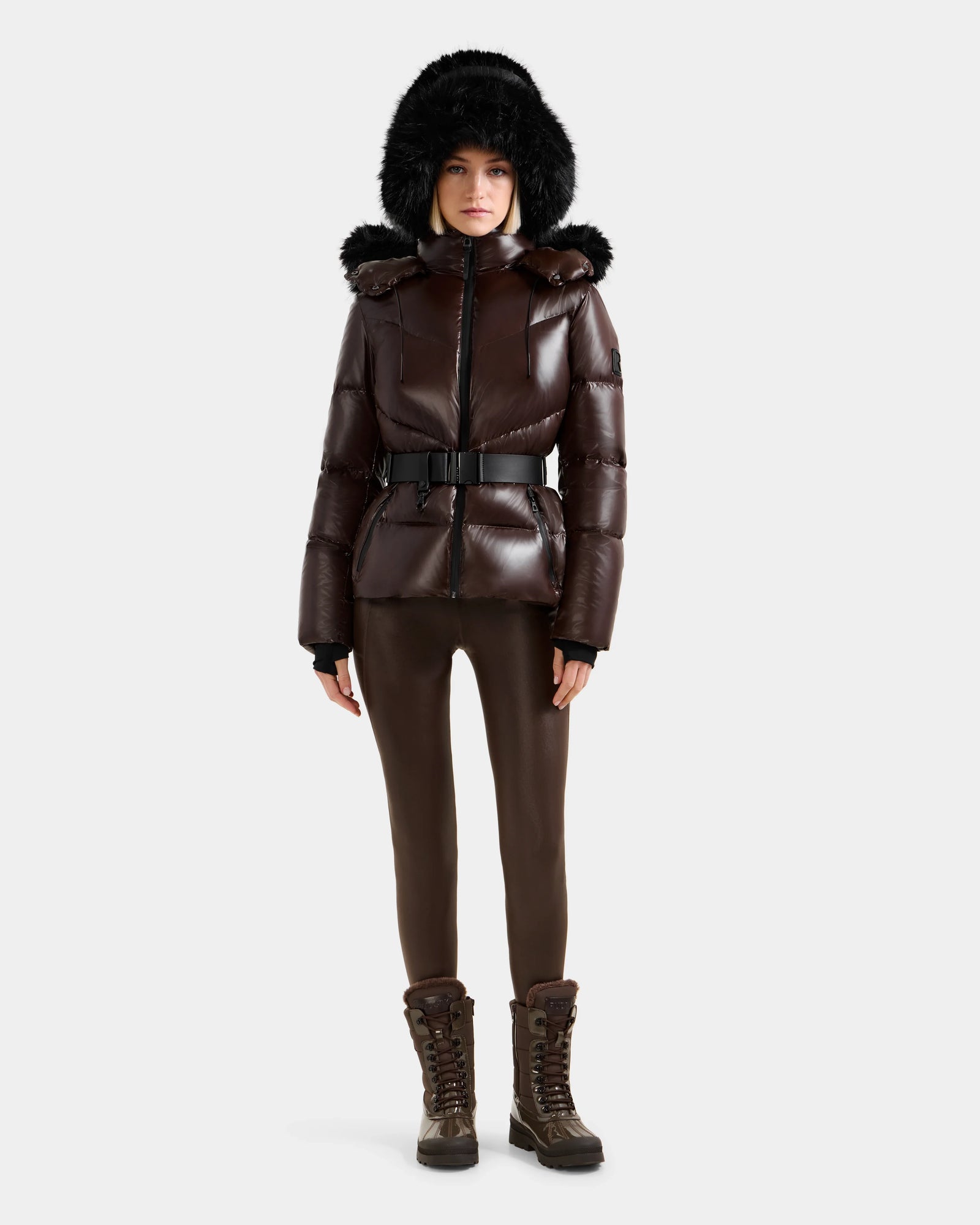 A woman wears the Rudsak CHRISTELLE S - L125544 Espresso down puffer jacket with faux fur trim hood, styled with brown leggings, a black belt, and brown lace-up boots, standing against a white background.