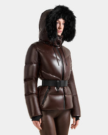 A person wears the Rudsak CHRISTELLE S - L125544 Espresso, a shiny brown women’s down puffer with black faux fur trim hood and black belt, paired with matching brown pants against a plain light background.