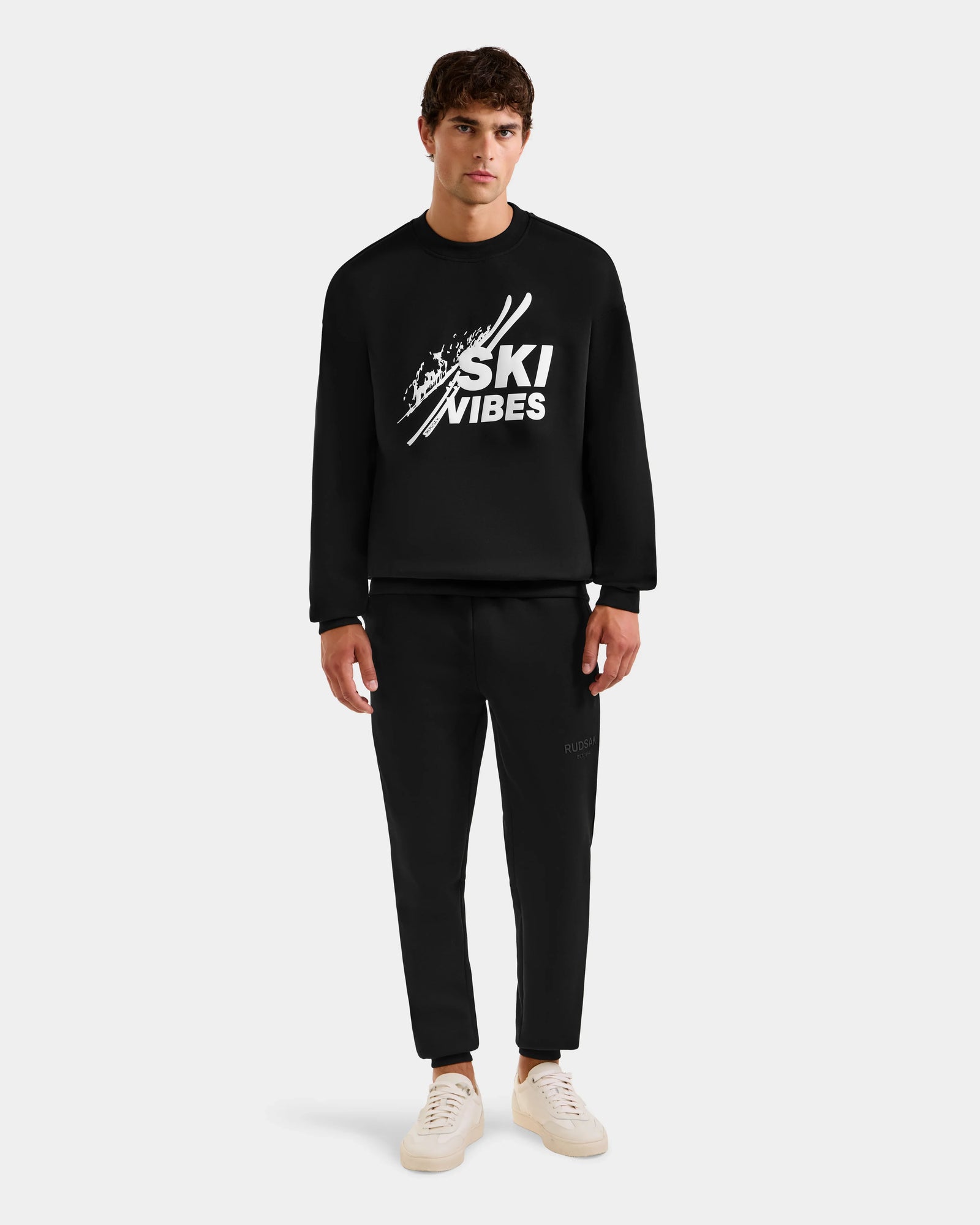 A man faces forward in the Rudsak COLSON - M525501 Black men’s crewneck sweatshirt with the SKI VIBES logo, matching black pants, and white sneakers, all against a plain light gray background. Made from premium cotton-blend fleece.