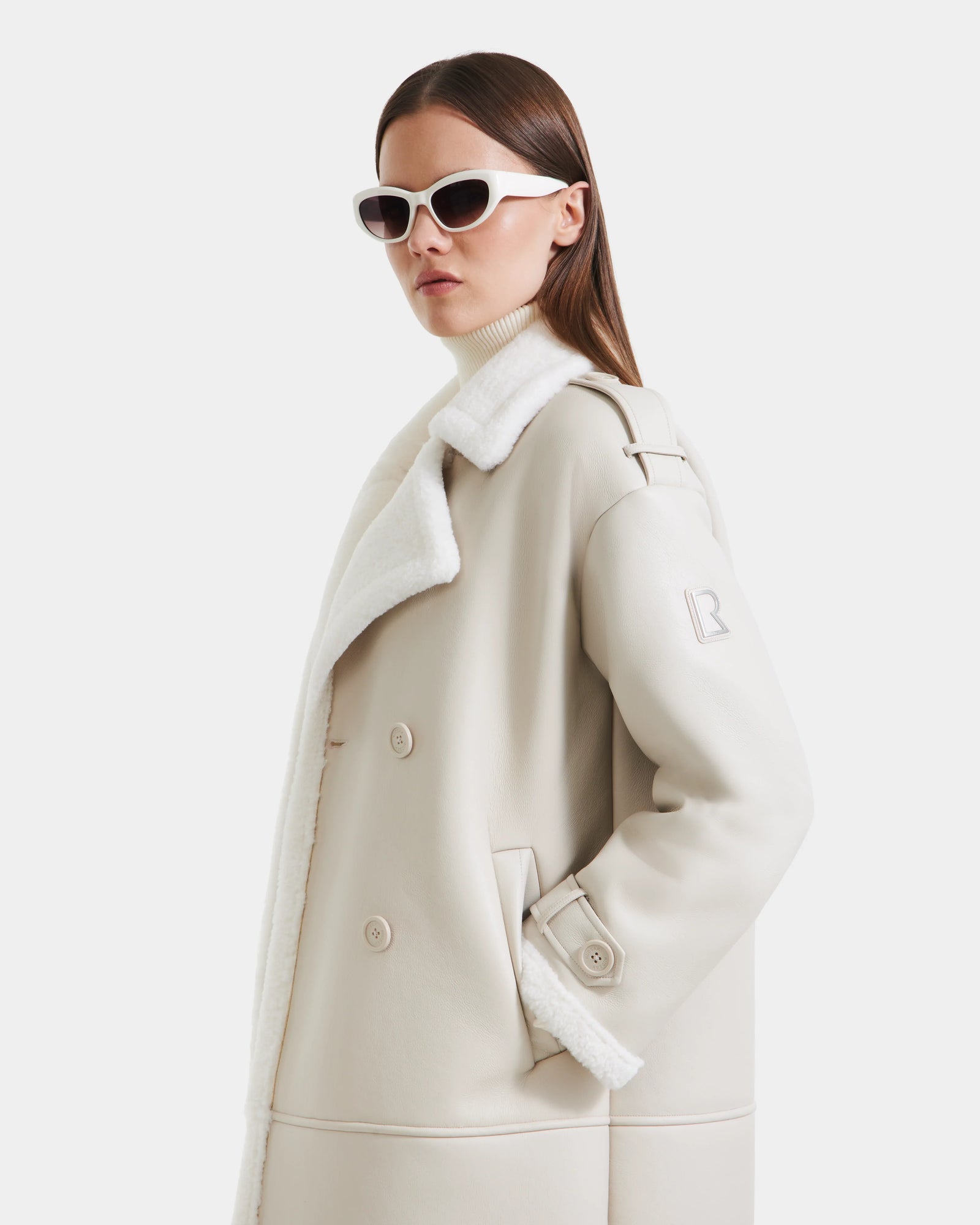 A woman in the Rudsak DARCY - L125539 Vanilla women's winter coat poses against a plain white background, wearing white sunglasses and a light turtleneck, looking confidently to the side.