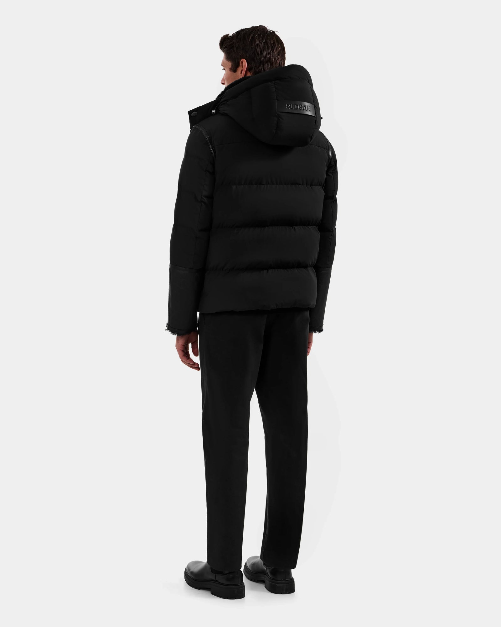 A man stands with his back to the camera, wearing the Rudsak DAVID - M125508 Black puffer jacket with a hood, paired with black pants and shoes, set against a plain light gray background.