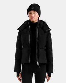 A person faces forward in a neutral pose, wearing the Rudsak DELILAH M - L125538 Black women’s down puffer with removable sleeves, paired with a black knit beanie and black pants, set against a plain light gray background.