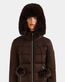 A person stands facing forward against a plain light background, wearing the Rudsak DEMI - L425539 Espresso women's faux-fur aviator hat with ear flaps and pom-pom ties, paired with a matching brown puffer jacket.