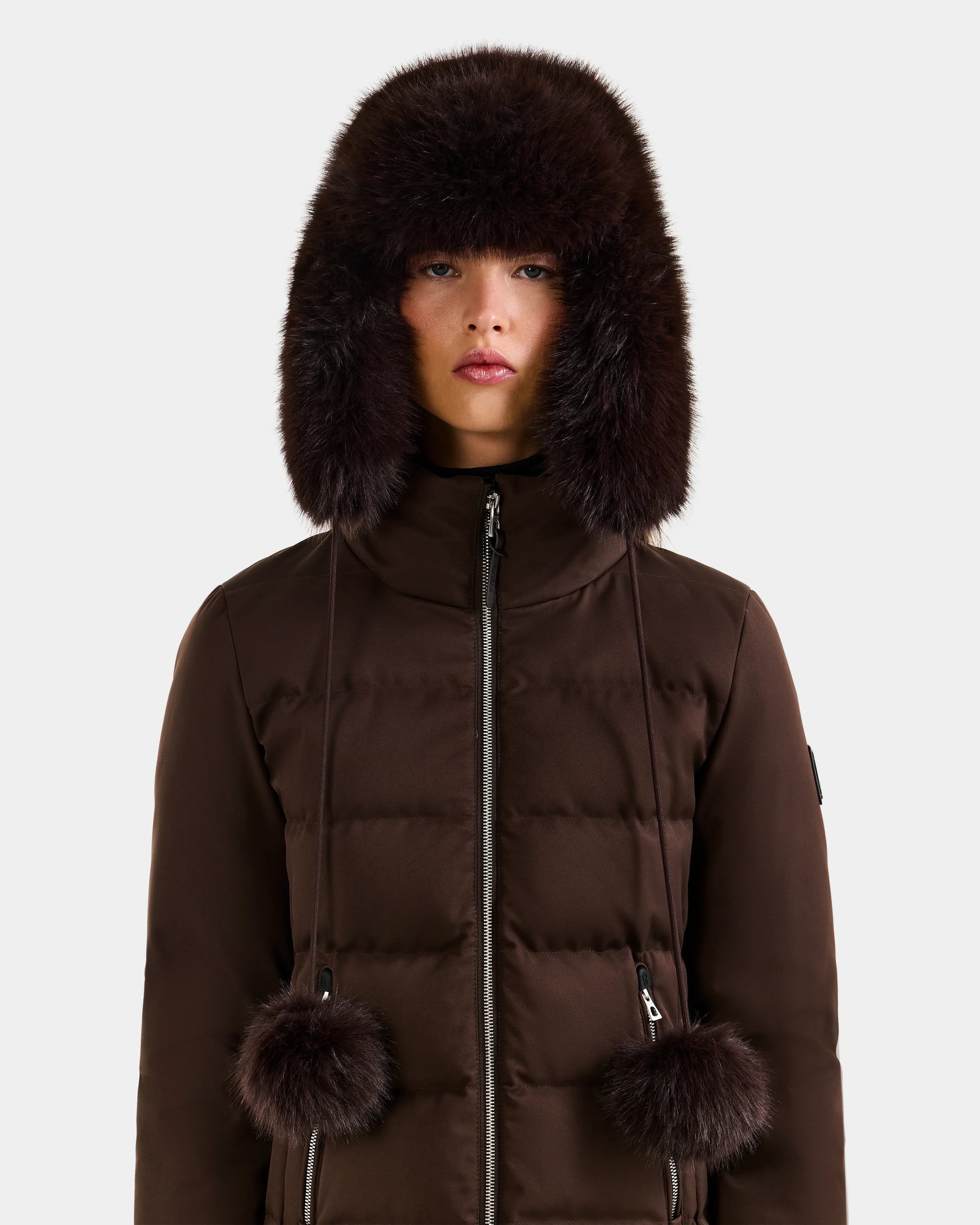 A person stands facing forward against a plain light background, wearing the Rudsak DEMI - L425539 Espresso women's faux-fur aviator hat with ear flaps and pom-pom ties, paired with a matching brown puffer jacket.