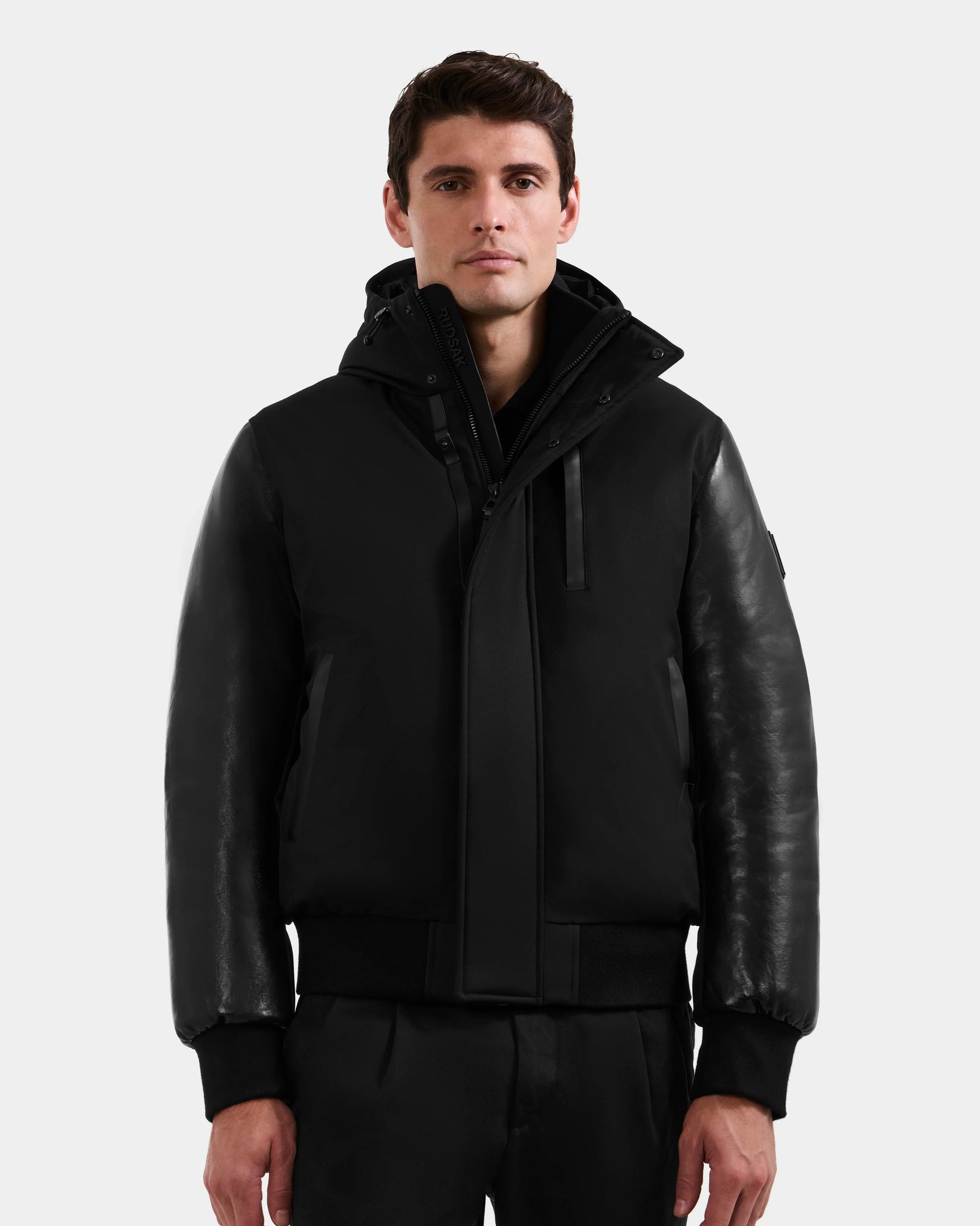 A man with short brown hair models Rudsak’s DERON - M125539 Black men’s down bomber, featuring vegan-leather sleeves and RDS-certified down, paired with black pants against a plain light gray background.