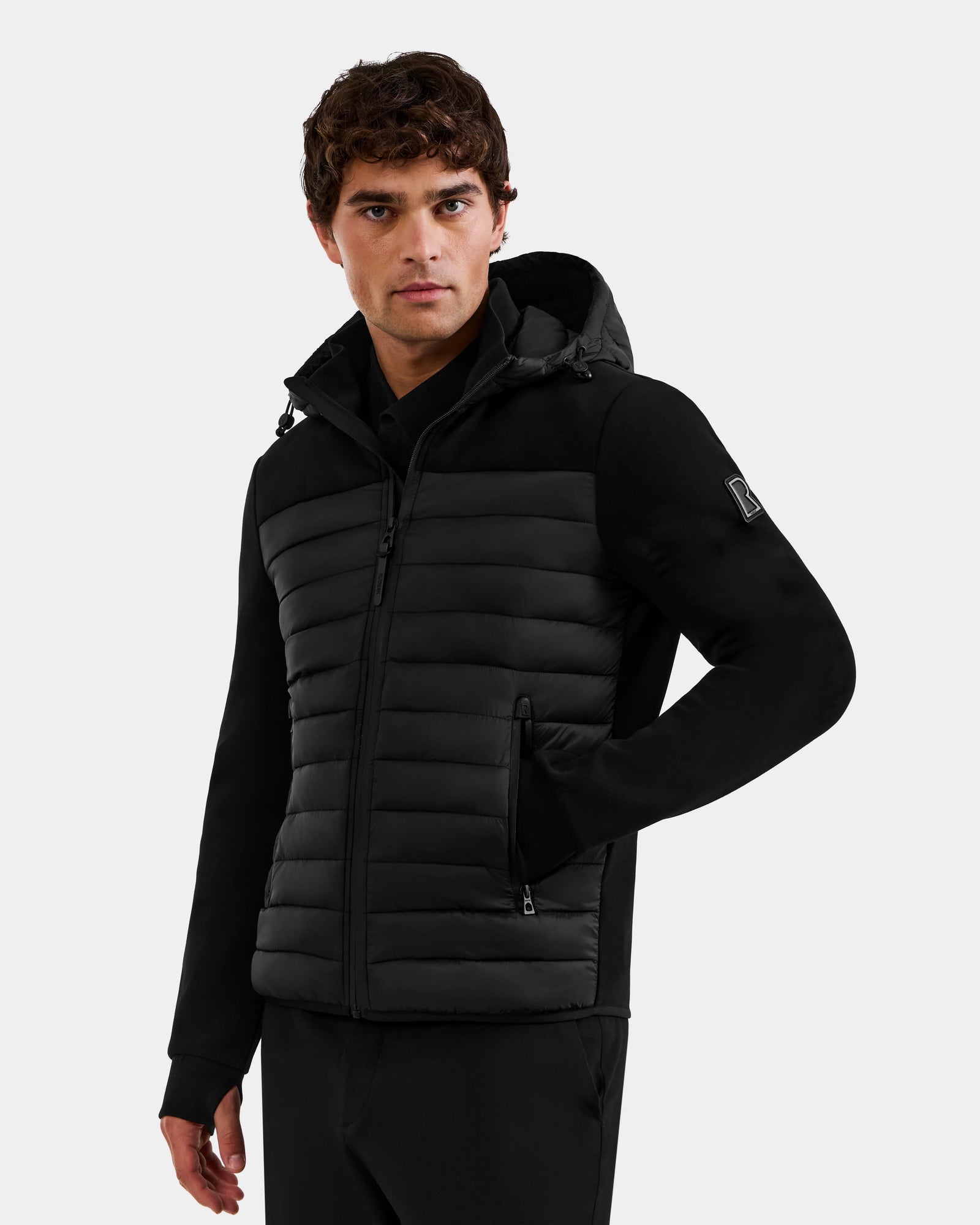 A man wearing the Rudsak DURIN M - M125530 Black hooded jacket, featuring lightweight down insulation, zippered pockets, and a sleeve patch, stands against a plain light gray background.