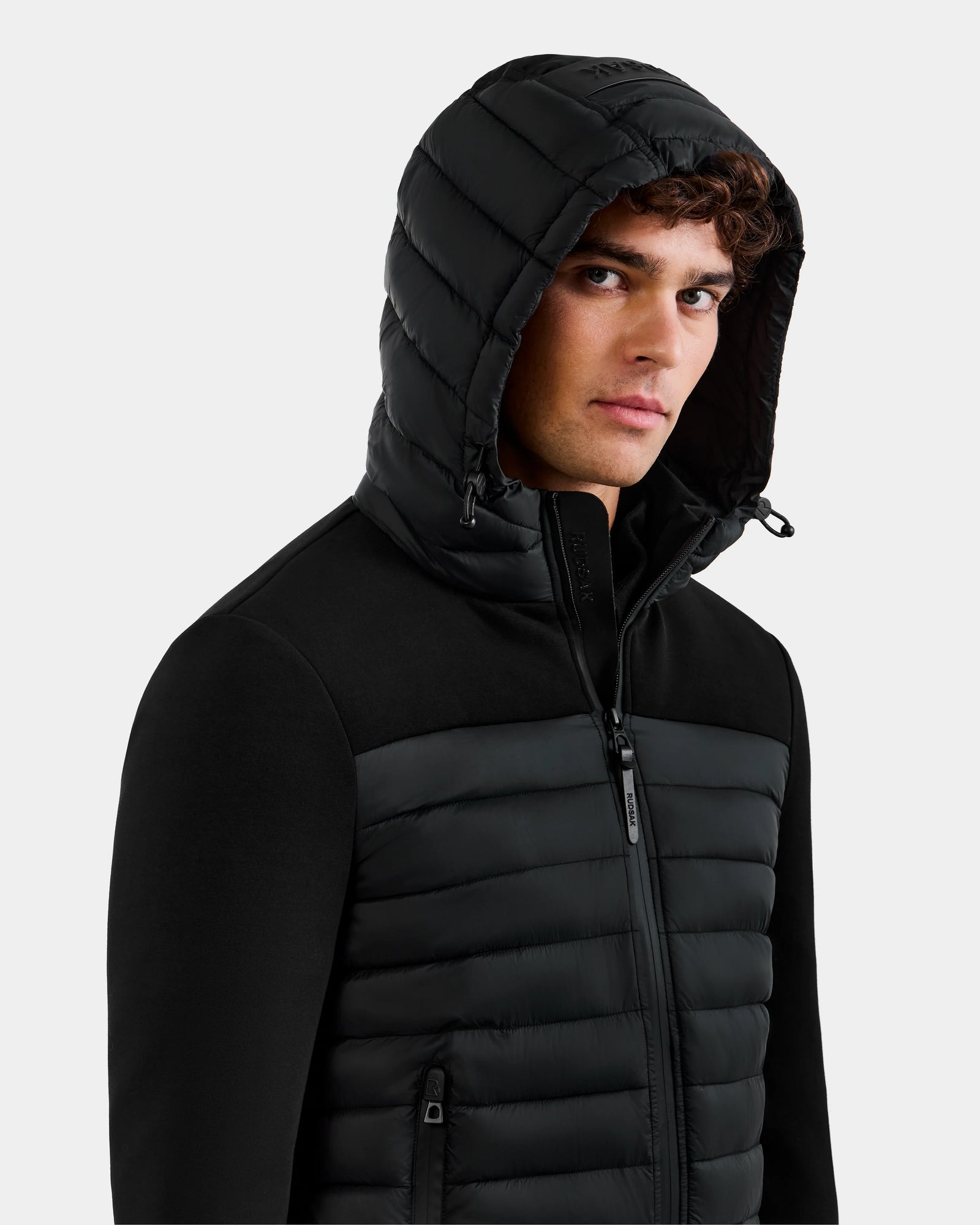 A young man in the DURIN M - M125530 Black men's hooded jacket by Rudsak, with lightweight down insulation, waterproof zippers, zip-up front, and side pockets, looks slightly to the side against a plain light background.