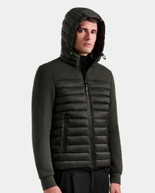 A young man in the Rudsak DURIN M - M125530 Dark Olive men’s hooded jacket, featuring lightweight down insulation and waterproof zippers, stands against a plain light background, looking slightly to the side with a neutral expression.