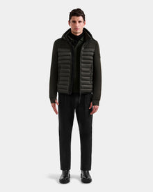 A man faces forward wearing the Rudsak DURIN M – M125530 Dark Olive men’s hooded jacket with lightweight down insulation and waterproof zippers, paired with black pants and boots, against a plain light gray background.