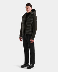 A man stands facing forward, wearing the Rudsak DURIN M - M125530 Dark Olive men's hooded jacket, paired with black pants, a black turtleneck, and black boots against a plain light background.