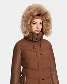 A woman in the Rudsak ELSA - L123535 Taupe quilted down puffer with leather sleeves, a high collar, and faux fur-trimmed hood pairs it with a matching knit hat against a light background—highlighting its sustainable design.