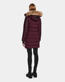 A woman wearing the Rudsak ELSA - L123535 Wine puffer coat with a faux fur-trimmed hood, black leggings, and boots stands facing away against a plain light background.