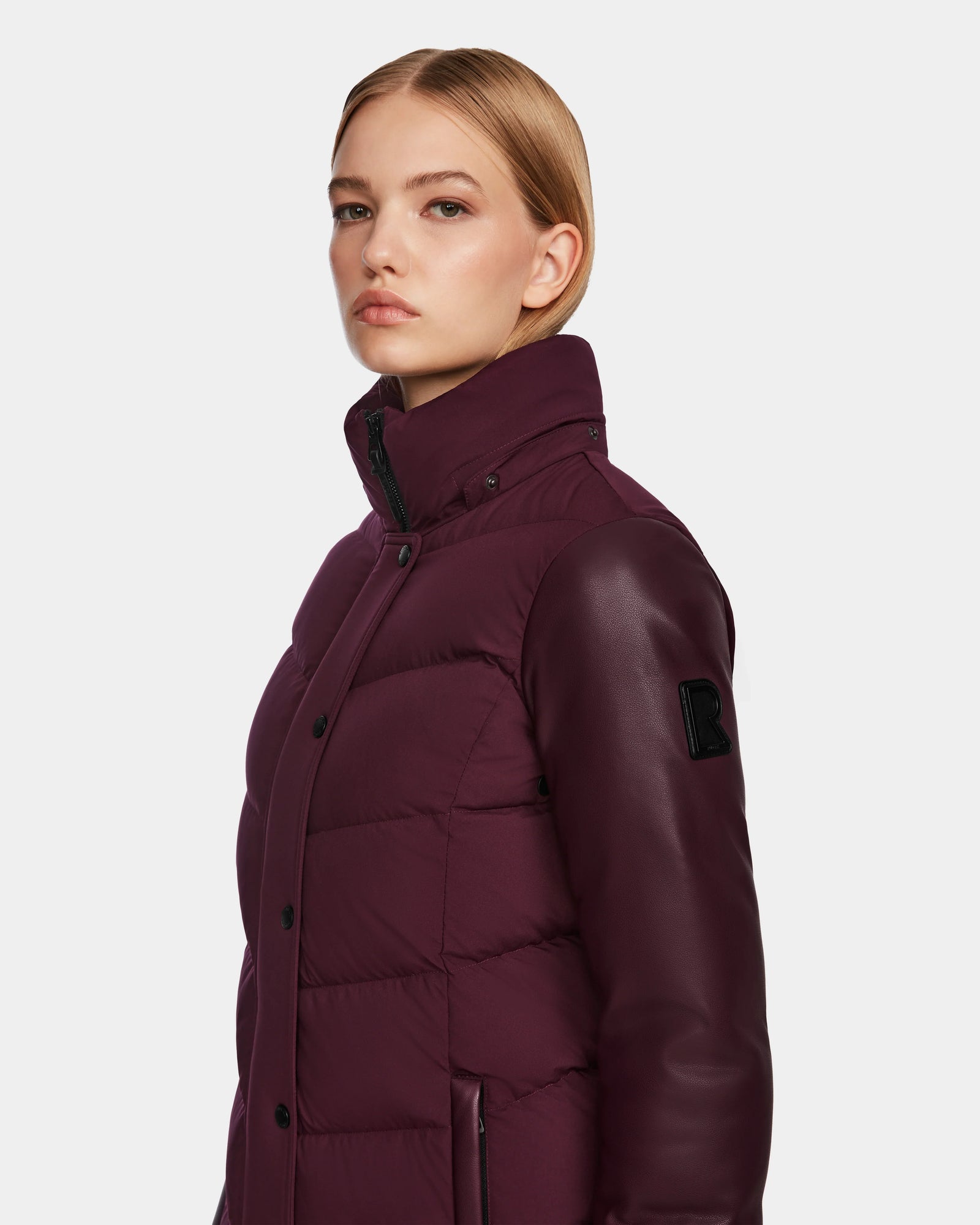 A woman with straight blonde hair models the Rudsak ELSA - L123535 Wine women’s quilted down puffer jacket, featuring leather sleeves and a high collar, posing confidently against a plain light background.