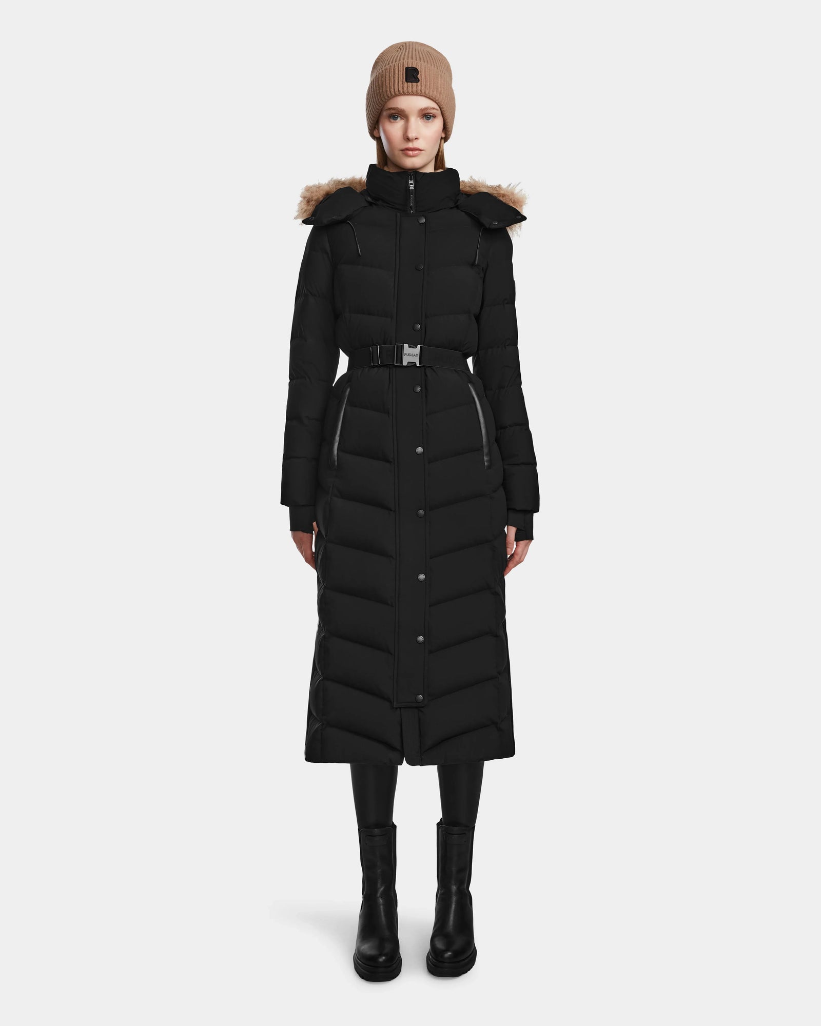A woman stands facing forward against a plain white background, wearing the Rudsak ELSIE M - L123517 Black/Natural maxi down puffer with belt and fur-trimmed hood, paired with black boots and a tan knit beanie.