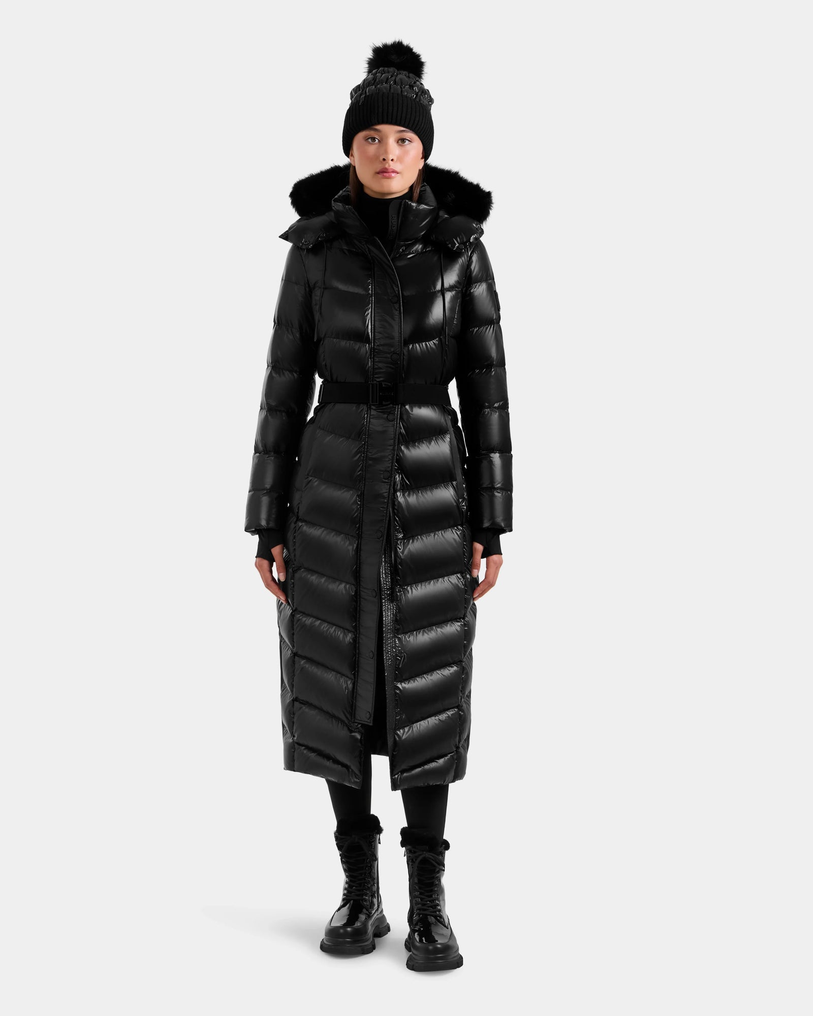 A person stands against a plain light background wearing the Rudsak ELSIE S - L124536 Black women’s shiny black, water-resistant maxi down puffer with fur-trimmed hood, paired with a black beanie with pom-pom, black boots, and pants.
