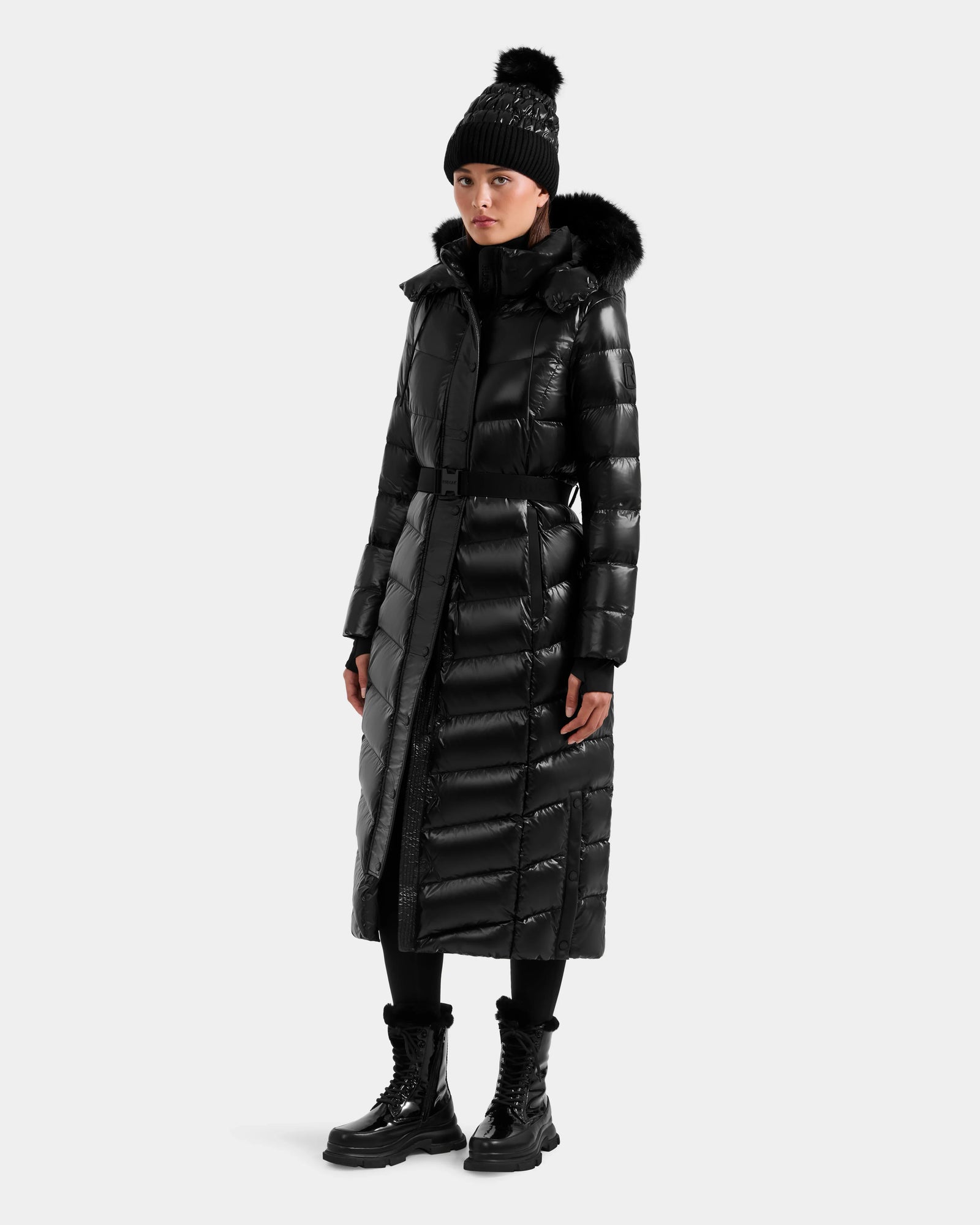 A person wearing the Rudsak ELSIE S - L124536 Black women’s maxi down puffer, paired with a black pom-pom beanie, boots, and leggings, stands against a plain light background.