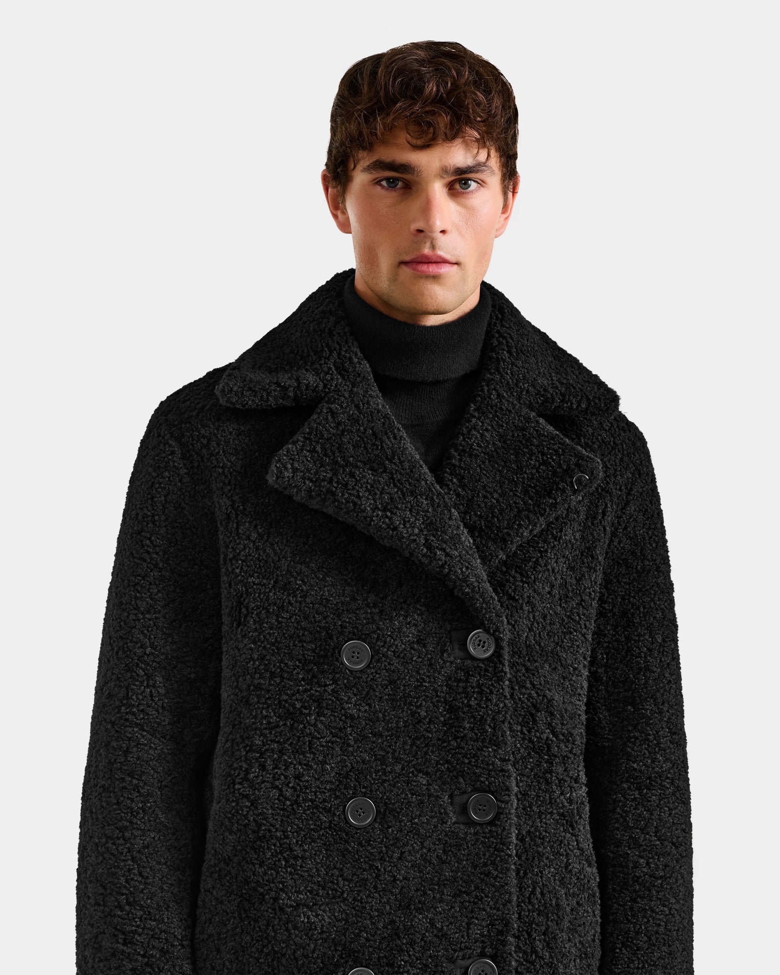 A man wearing the Rudsak ELTON - M125534 Black double-breasted overcoat over a black turtleneck stands against a plain light gray background, looking directly at the camera.