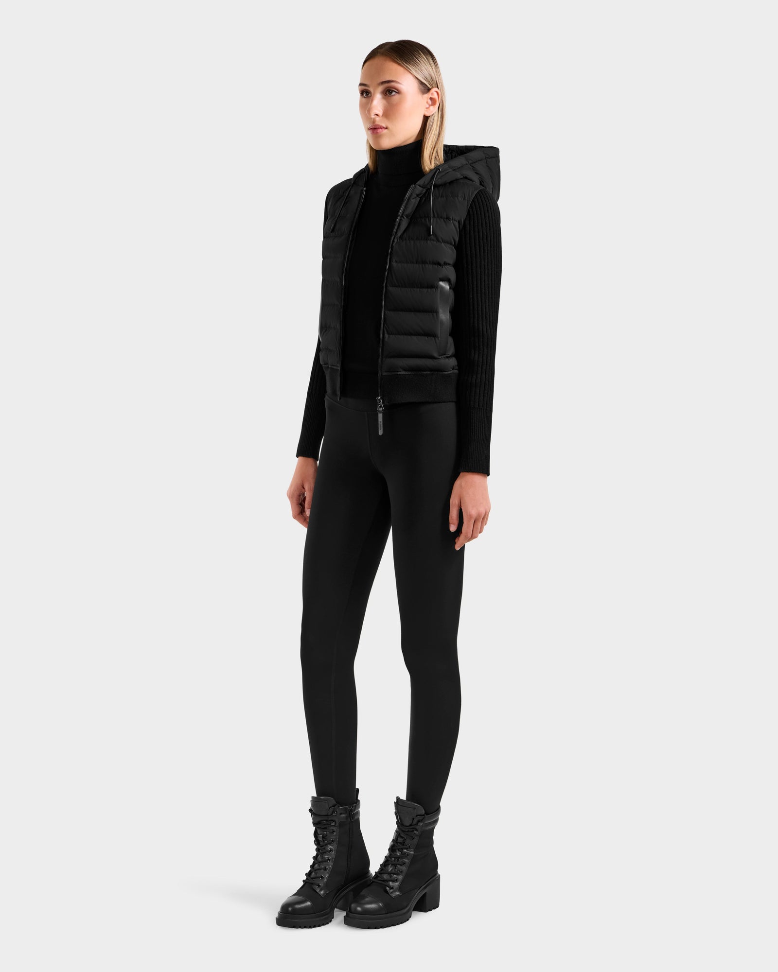 A woman wears the Rudsak EMILY - L125515 Black women’s hooded down bomber with RDS-certified down, a black turtleneck, leggings, and lace-up boots, standing against a plain light gray background with arms relaxed at her sides.