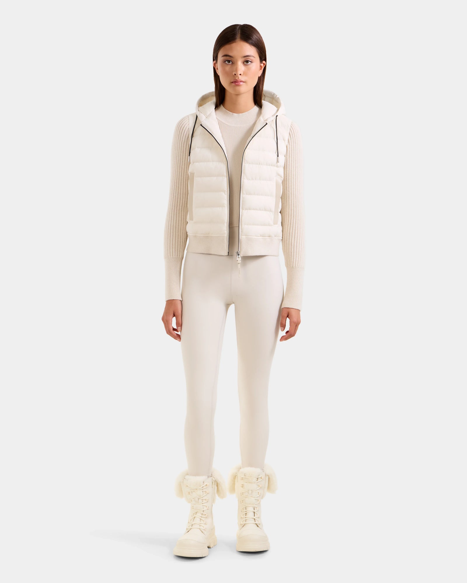 A woman faces forward in a vanilla Rudsak EMILY - L125515 women’s hooded down bomber vest, ribbed long-sleeve top, fitted leggings, and fur-trimmed boots against a plain light gray background.