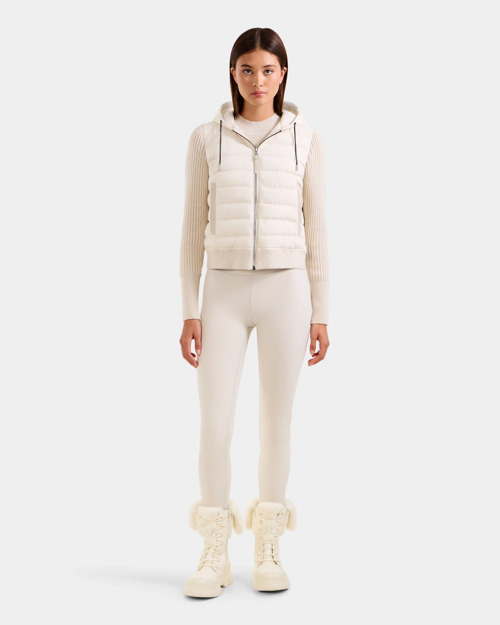 A woman stands facing forward wearing the Rudsak EMILY - L125515 Vanilla hooded down bomber over a cream ribbed sweater and matching leggings with fuzzy white lace-up boots, set against a plain light background.