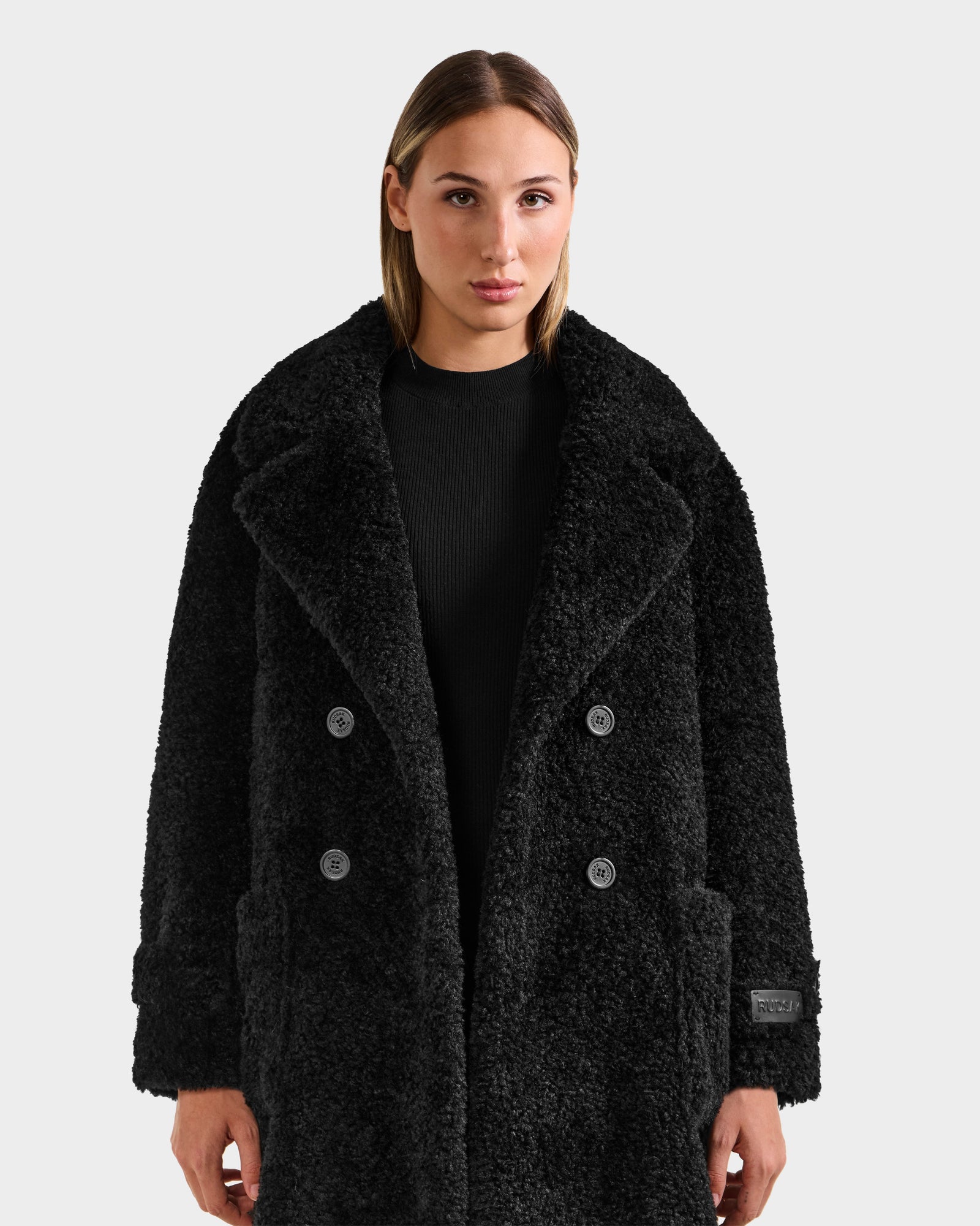 A woman with straight, light brown hair wears the Rudsak EMRYN - L125588 Black teddy jacket over a black shirt, standing against a plain light gray background and looking directly at the camera.