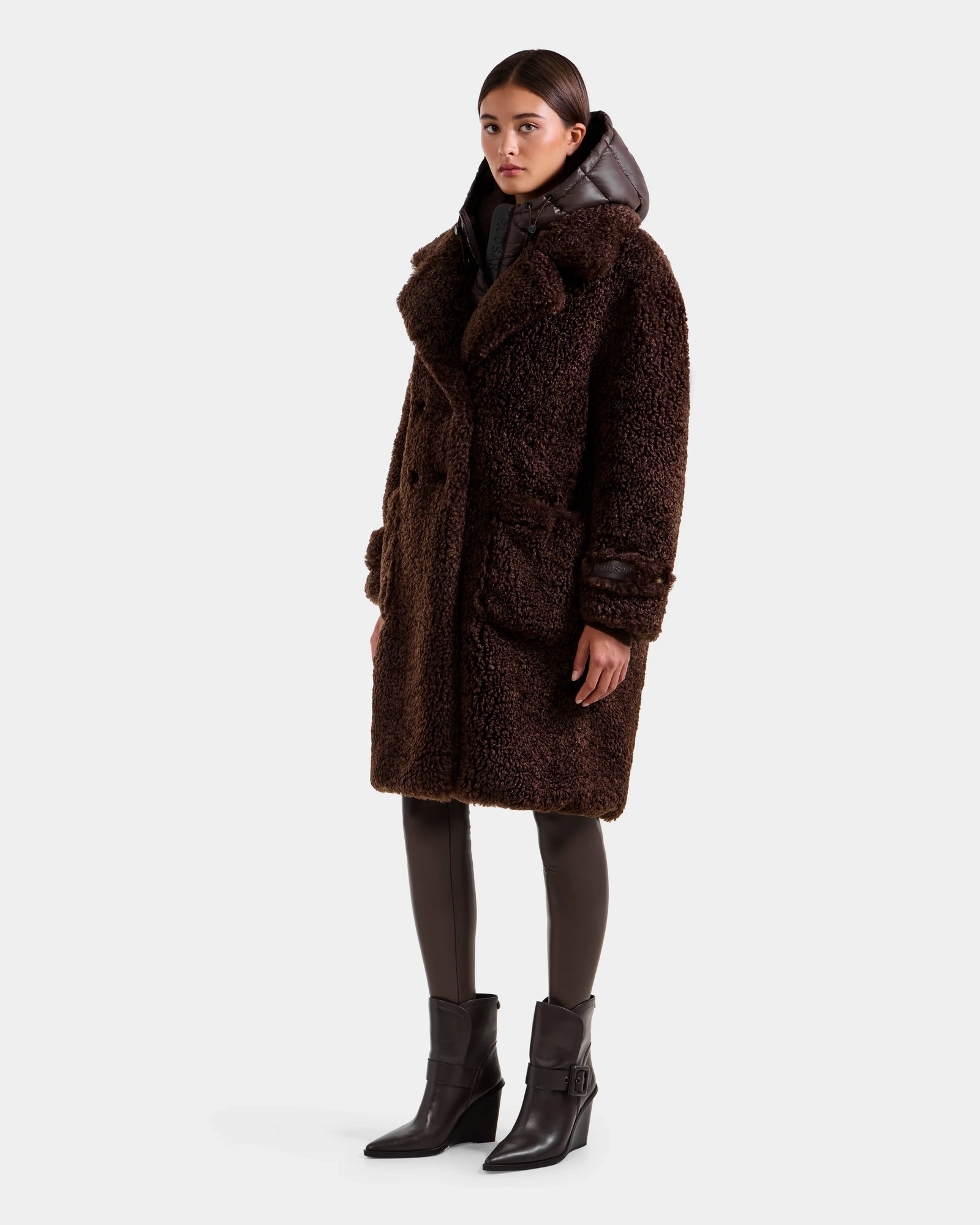 A woman wears the Rudsak EMRYN - L125588 Espresso textured teddy jacket as a knee-length winter coat over a black puffer, paired with dark pants and heeled ankle boots, standing against a light gray background.